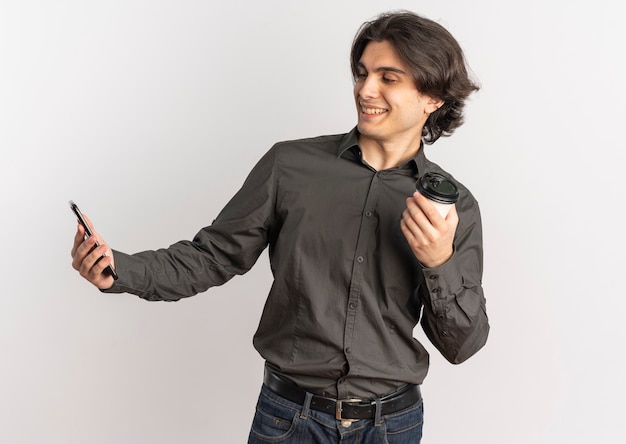 Young pleased handsome caucasian man holds phone and coffee cup looking at phone isolated on white background with copy space