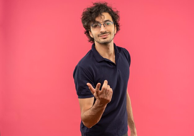 Young pleased man in black shirt with optical glasses holds hand out isolated on pink wall