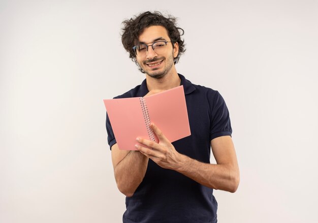 Young pleased man in black shirt with optical glasses holds and looks at notebook isolated on white wall