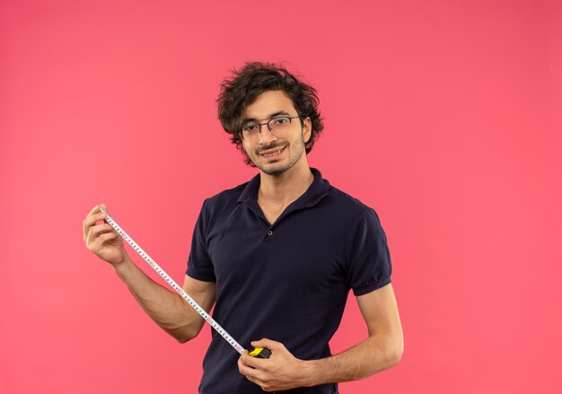 Il giovane uomo soddisfatto in camicia nera con vetri ottici tiene il metro a nastro isolato sulla parete rosa