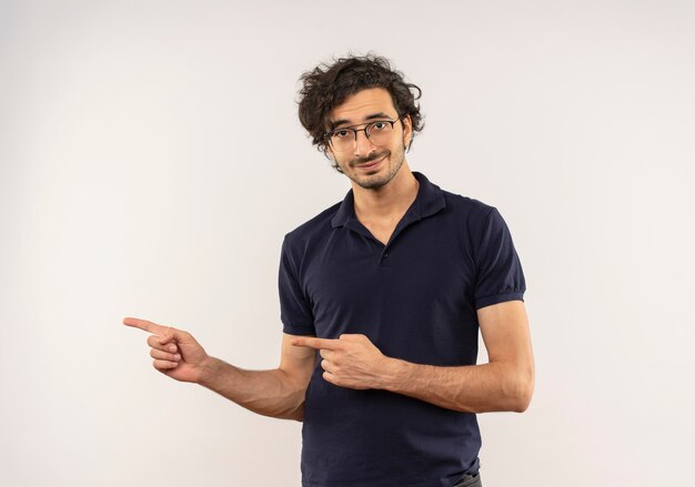 Young pleased man in black shirt with optical glasses points at side and looks isolated on white wall