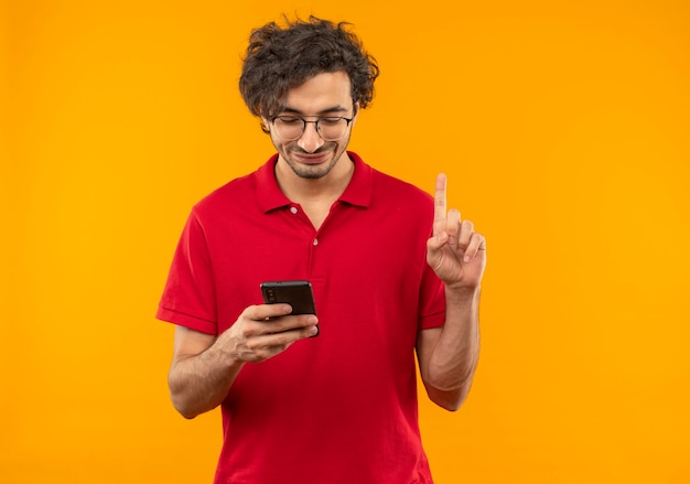 Young pleased man in red shirt with optical glasses holds phone and points up isolated on orange wall