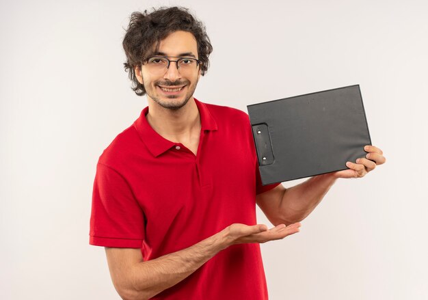 Young pleased man in red shirt with optical glasses holds and points at clipboard isolated on white wall