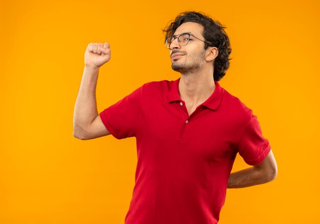 Young pleased man in red shirt with optical glasses raises fist up isolated on orange wall