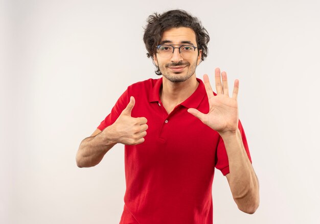 Young pleased man in red shirt with optical glasses thumbs up and raises hand isolated on white wall