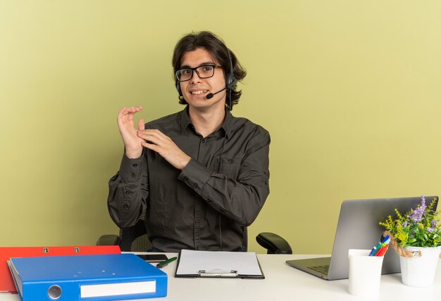 Young pleased office worker man on headphones in optical glasses sits at desk with office tools using laptop holds hands together isolated on green background with copy space