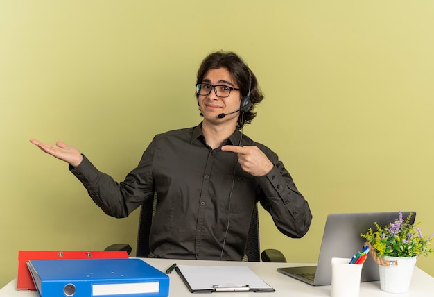 Young pleased office worker man on headphones in optical glasses sits at desk with office tools using laptop points at empty hand