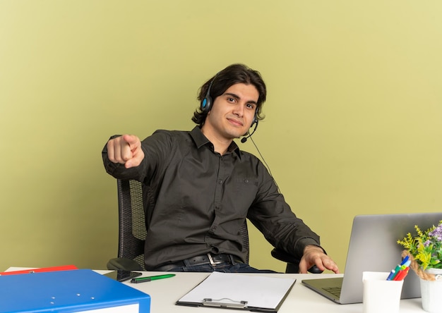 Uomo di lavoratore di ufficio felice giovane sulle cuffie si siede alla scrivania con strumenti di ufficio utilizzando laptop che punta alla telecamera isolata su sfondo verde con spazio di copia