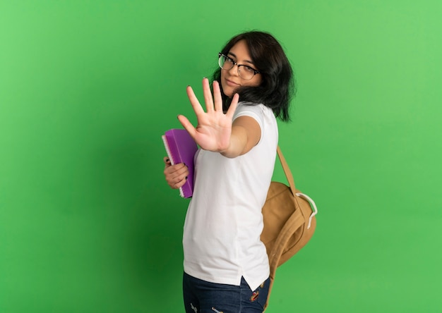 Молодой доволен довольно кавказской школьницей в очках и жестами на спине, стоп знак рукой, держащий книги, изолированные на зеленом пространстве с копией пространства