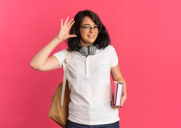 Молодая довольная симпатичная кавказская школьница в сумке для очков и наушниках на шее держит книгу и тетрадь с поднятой рукой на розовом с копией пространства