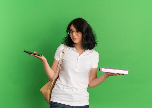 Young pleased pretty caucasian schoolgirl wearing glasses and back bag holds book and phone on green  with copy space