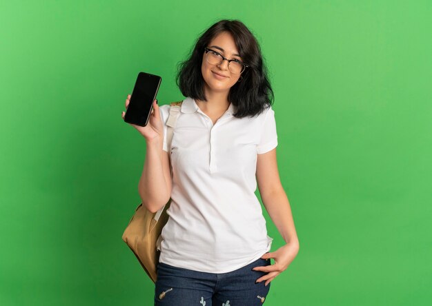 Молодая довольная симпатичная кавказская школьница в очках и задней сумке держит телефон на зеленом с копией пространства