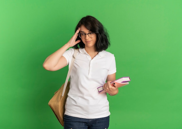 Young pleased pretty caucasian schoolgirl wearing glasses and back bag puts hand on forehead looking down holding books on green  with copy space