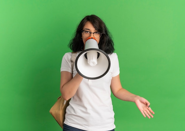 Young pleased pretty caucasian schoolgirl wearing glasses and back bag speaks through loud speaker on green  with copy space