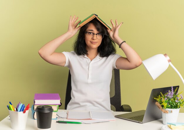 Young pleased pretty caucasian schoolgirl wearing glasses sits at desk with school tools holds book over head on green  with copy space