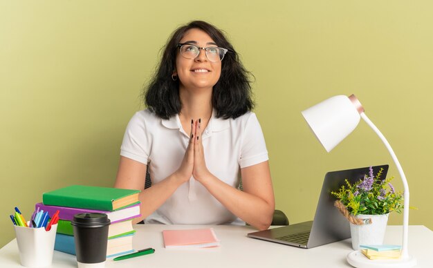 Young pleased pretty caucasian schoolgirl wearing glasses sits at desk with school tools looks up holding hands together isolated on green space with copy space