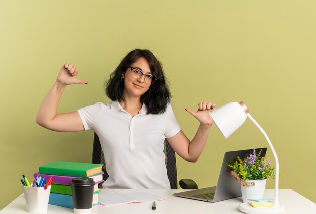 Young pleased pretty caucasian schoolgirl wearing glasses sits at desk with school tools points at herself isolated on green space with copy space