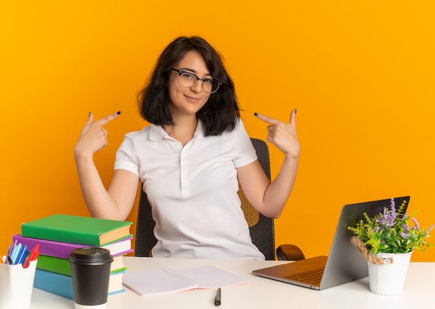 Young pleased pretty caucasian schoolgirl wearing glasses sits at desk with school tools points at herself on orange  with copy space