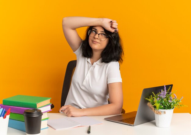 Young pleased pretty caucasian schoolgirl wearing glasses sits at desk with school tools puts arm on forehead looking at side on orange  with copy space