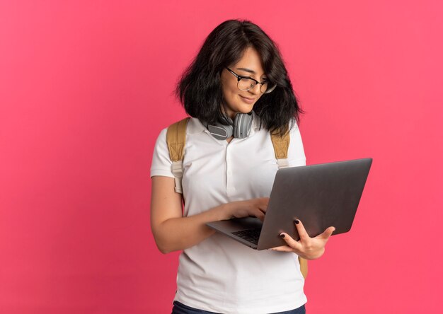 Young pleased pretty caucasian schoolgirl with headphones on neck wearing glasses and back bag holds and looks at laptop on pink  with copy space