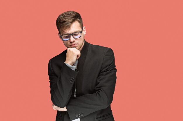 Young pondering handsome man in black suit and glasses isolated on red studio background