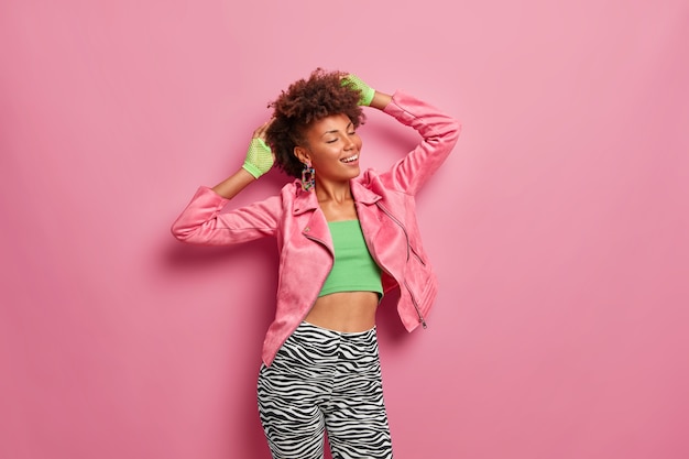 Young positive Afro American woman in good physical shape, stands gladfully indoor, smiles sincerely, closes eyes with pleasure, dressed in stylish colorful outfit