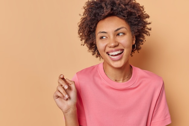 Young positive beautiful woman with curly hair looks happily away feels good has carefree expression wears casual t shirt isolated over brown backgrounnd. Human emotions and feelings concept