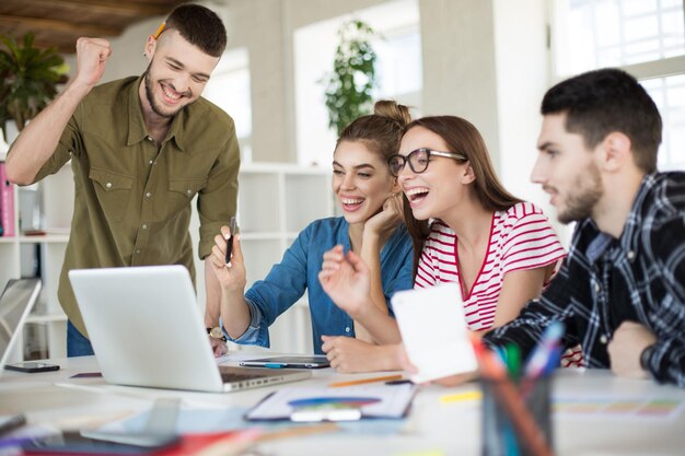 Молодые позитивные деловые люди счастливо работают на ноутбуке вместе Группа улыбающихся мужчин и женщин смеется, проводя время на работе в современном уютном офисе