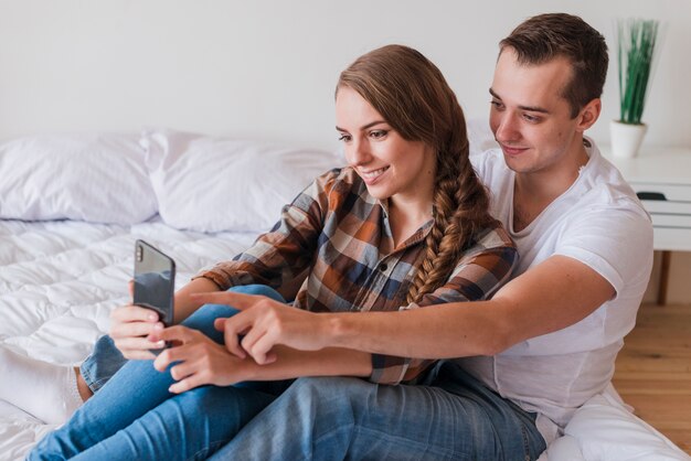 Молодая позитивная пара с помощью смартфона в спальне у себя дома