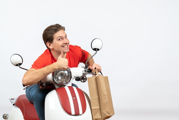 Foto gratuita ragazzo giovane corriere positivo in uniforme rossa che si siede sul motorino che tiene gli ordini e che fa gesto giusto sulla parete gialla