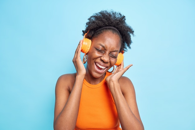 Young positive curly Afro American woman enjoys listening music in wireless stereo headphones