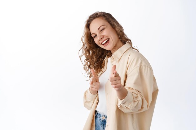 Young positive girl winking and smiling at you pointing fingers camera praising or inviting people standing over white background Copy space