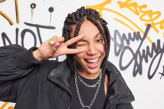 Free photo young positive graffiti artist makes peace gesture over eye smiles broadly dressed in black jacket and metal chians around neck poses against colorful drawn street wall