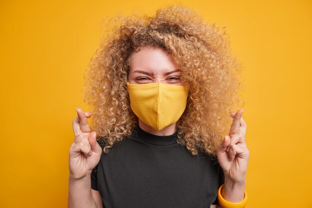 Young positive woman has fair curly hair crosses fingers anticipates good positive results hopes dreams come true wears black t shirt and disposable mask to prevent virus spread isolated on yellow