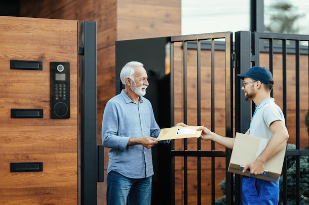 Giovane impiegato delle poste che effettua la consegna a domicilio e dà un pacco al cliente senior al cancello d'ingresso