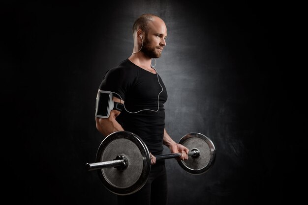 Young powerful sportsman in heaphones training with barbell over black wall