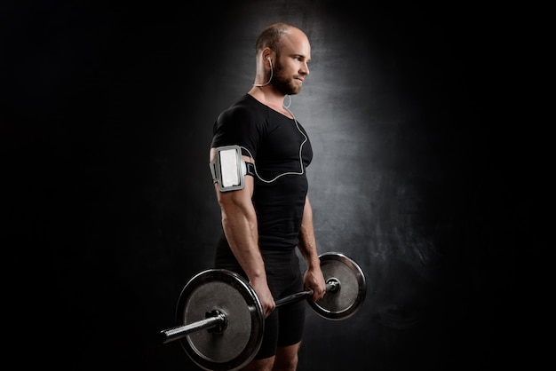 Young powerful sportsman training with barbell over black wall.