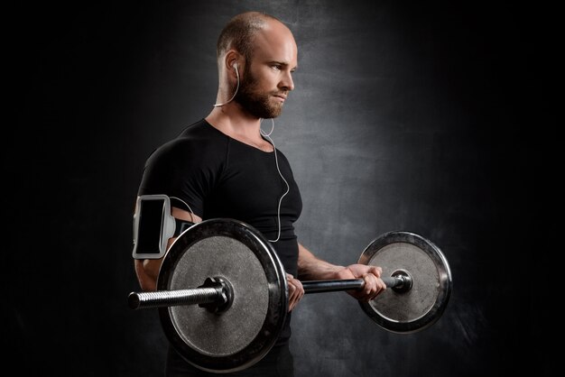 Young powerful sportsman training with barbell over black wall.