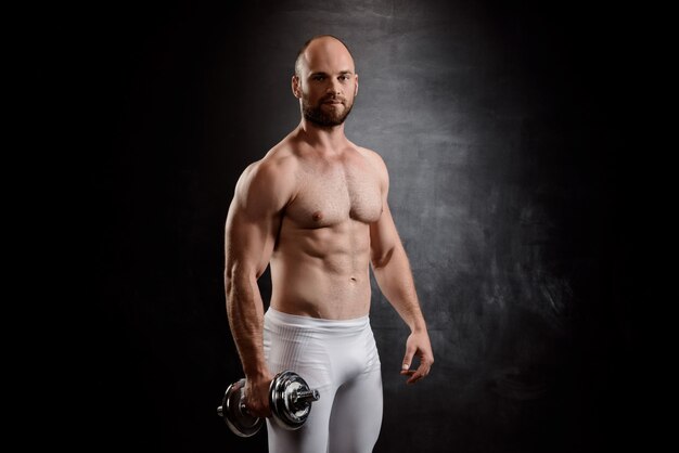 Young powerful sportsman training with dumbbells over black wall.