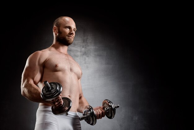 Young powerful sportsman training with dumbbells over black wall.