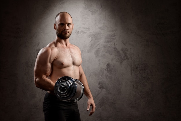 Young powerful sportsman training with dumbbells over dark wall.