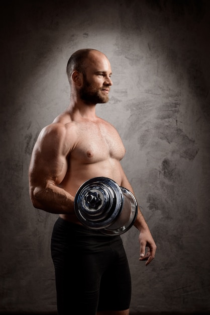 Young powerful sportsman training with dumbbells over dark wall.