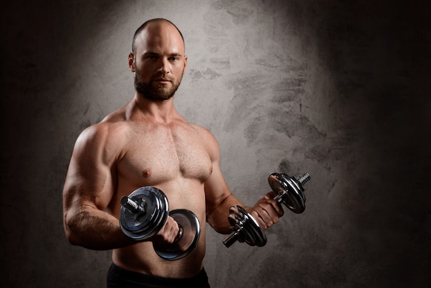 Young powerful sportsman training with dumbbells over dark wall.