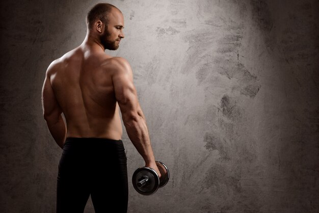 Young powerful sportsman training with dumbbells over dark wall.