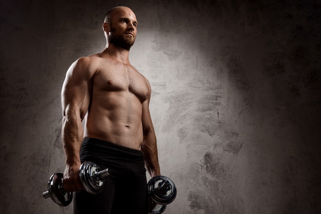 Young powerful sportsman training with dumbbells over dark wall.