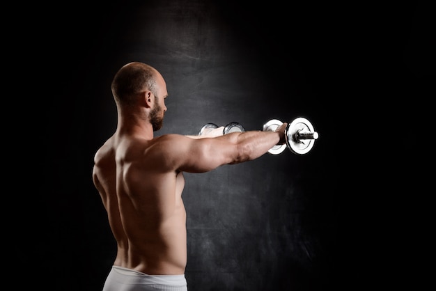 Бесплатное фото Тренировка молодого мощного спортсмена с гантелями над черной стеной.