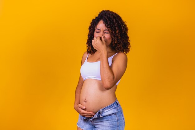 Young pregnant afro woman standing on yellow studio background, smelling something stinky and disgusting, intolerable smell, holding breath with fingers on nose. bad smell