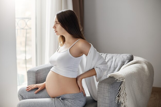 Young pregnant female rubbing her back to relieve pains. Staying in her warm and cozy home.