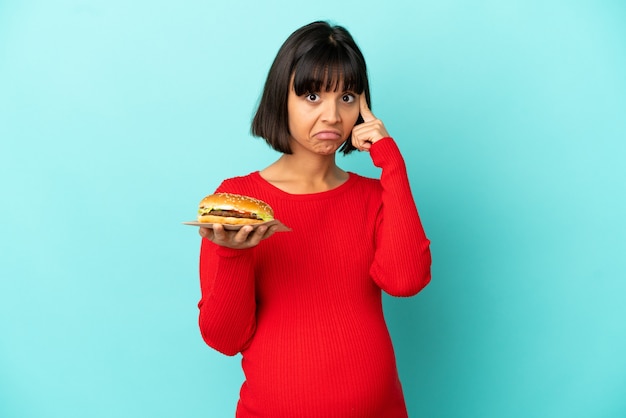 Молодая беременная женщина, держащая гамбургер на изолированном фоне, думает об идее