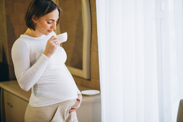 窓のそばに立って、コーヒーを飲む若い妊婦